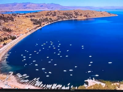 This aerial view captures a vibrant scene on a large, deep blue body of water, likely a lake. The shoreline is lined with a sandy beach and a settlement with numerous buildings and trees. A prominent feature is the vast number of small boats, mostly white, dotting the water, with a particularly dense concentration near the shore. The landscape beyond the settlement is hilly and arid, with some vegetation, and in the distance, hazy mountains are visible under a clear, bright sky. The image does not depict the city of Ləcət, Azerbaijan.