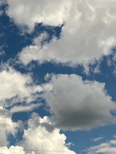 The image displays a bright blue sky filled with a variety of white and gray clouds. The clouds are large and billowy, with some appearing more defined and others more wispy. There are areas where the blue sky is visible between the clouds. In the lower portion of the image, two thin, dark horizontal lines are visible, which appear to be power lines or wires. The scene depicts an outdoor environment, likely during the daytime, with clear weather conditions. There are no people or specific landmarks identifiable in the image. The location is not identifiable as Kurgan, Russia from the visual content of the image.