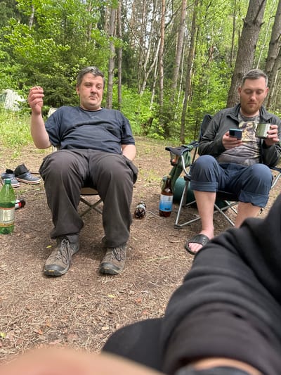 The image depicts a scene in what appears to be a forest clearing in Папярнянскі сельскі Савет, Belarus. Two men are present. One man is sitting in a folding chair, wearing a dark t-shirt, dark trousers, and hiking boots. He is turned towards the camera and is holding a cigarette in his hand. Several empty bottles, including a large green plastic bottle and smaller glass beer bottles, are at his feet. Another man is visible in the background, also sitting in a folding chair. He is wearing shorts, a t-shirt, and a jacket. He is holding a cell phone in one hand and a metal mug in the other. There are trees and green foliage surrounding the clearing. The ground is covered in dry leaves and pine needles.
