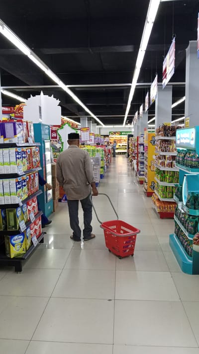 A man, wearing a light brown long-sleeved shirt, dark pants, and a peci, is pushing a red shopping basket with wheels down a supermarket aisle. The aisle is lined with shelves stocked with various products. In the foreground, to the left of the man, there are shelves filled with boxed items, some with colorful packaging. One box prominently displays the brand "Nutrilon Royal." To the right of the man, the aisle continues with more shelves containing products in various packaging, including what appear to be snacks and other groceries. In the background, the aisle extends further, with more shelves and shoppers visible. Fluorescent lights are illuminating the scene from the black ceiling above. Signs are hanging from the ceiling, some with text, and others are decorative displays. One sign reads "KELUAR," which means "EXIT" in Indonesian. The overall setting is a well-lit, modern supermarket. The time of day is indeterminate, but it appears to be during business hours. The scene is factual and observational, with no discernible emotions conveyed by the subjects.