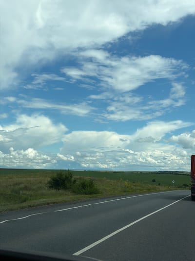The image shows a roadside view from a vehicle, likely a truck, driving on a paved road in Ostrovnoye, Russia. The perspective is from inside the vehicle, looking out. The scene is divided horizontally by the road which occupies the right portion of the image and a vast sky filled with clouds on the left.
The sky is a dominant feature, painted with a brilliant blue and adorned with a variety of clouds. Some are wispy and stretched, hinting at movement, while others are larger and more voluminous, suggesting fair weather or possibly developing cumulus formations.
Adjacent to the road is a grassy verge with some scattered yellow wildflowers and a couple of small bushes. Beyond this, stretching towards the horizon, is a large, green field, possibly farmland. The landscape is flat and appears to be rural.
In the far distance, the horizon is marked by a thin line of trees and a hazy, lighter blue sky. A road sign with a circular red border and a white symbol inside is visible on the left side of the road, near the field. The overall impression is one of a daytime journey through the countryside. The lighting suggests it's likely midday or early afternoon. There is no text visible in the image.