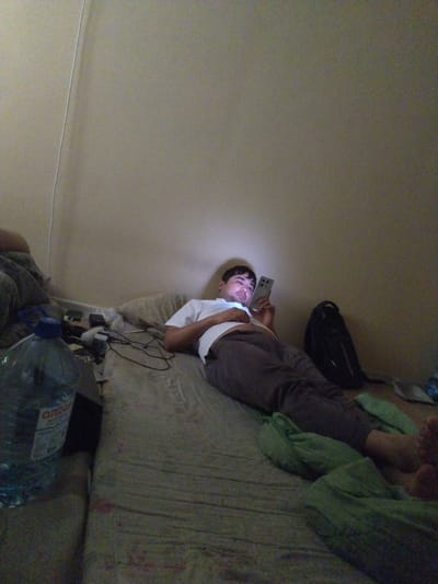 A man lies on his back on a narrow bed, illuminated by the screen of a smartphone he holds in his right hand. He is wearing a white t-shirt and dark grey sweatpants. The bed is covered with a light-colored, textured sheet and a green blanket. To the left of the bed is a large blue plastic water bottle with a blue cap, and a black bag containing electronics. Behind the man, a black backpack rests against the wall. The room appears dimly lit, with a plain beige wall forming the backdrop. The man's feet are visible at the bottom right of the frame, partially covered by the green blanket. The scene suggests a domestic or personal space within a building.