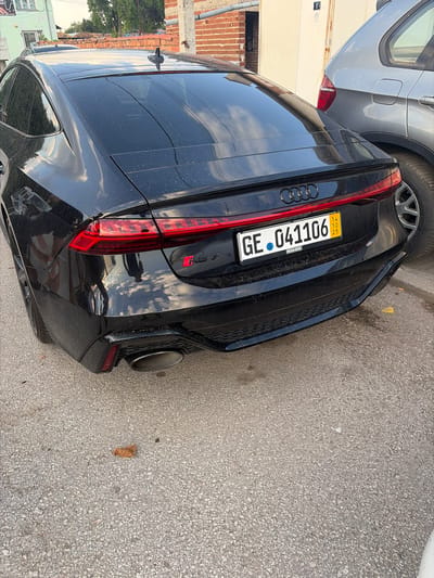 This media file shows the rear right side of a black Audi RS7 parked on a paved road. The car is in focus, with a portion of a silver car visible to its left. The scene appears to be outdoors during the daytime, with visible reflections of the sky and surrounding environment on the car's surface. The road is made of asphalt with visible gravel. There is some dust on the car's exterior, suggesting it may not have been recently washed.

The license plate of the Audi is clearly visible and reads "GE 041106". Above the license plate, the Audi rings emblem is visible on the car's trunk. The "RS 7" badging is also present on the rear of the car. The car has a prominent exhaust pipe on the right side. In the background, there are some buildings, a brick wall, and trees, suggesting an urban or semi-urban setting. The lighting suggests it is daytime, possibly late afternoon given the angle of the reflections. There are no people visible in the image. The overall impression is of a parked car in a functional, everyday environment.