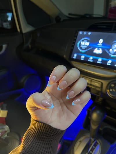 A person's hands are shown inside a vehicle, with a dashboard displaying a digital clock showing 18:58. The nails are manicured with a French tip design, featuring a light blue color on the tips. The interior of the car is illuminated with blue ambient lighting. The hand wears a brown, ribbed sweater. The background indicates the setting is likely an urban environment, as suggested by the possibility of being in the city of Beuringen, Indonesia.