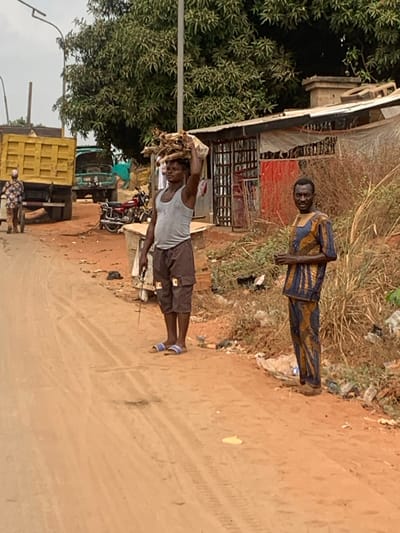 The scene in Aguleri, Nigeria, depicts an unpaved reddish-brown dirt road with scattered debris and tire tracks. On the left, a large yellow dump truck is parked, with a second green truck visible behind it. A male individual wearing a plaid shirt and dark trousers stands near the rear of the yellow truck, facing away. In the foreground, two adult males are positioned on the right side of the road. The man on the left wears a grey sleeveless tank top, dark cargo shorts, and blue-and-white slide sandals; he carries a bundle of light-colored sticks or firewood on his head, secured by a white fabric, and holds a thin object with a wire in his left hand. The man on the right wears a patterned blue, brown, and yellow long-sleeved tunic and matching trousers, and dark shoes; he stands facing the viewer with his hands held in front of him. Behind these two individuals are rudimentary structures with corrugated metal roofs and walls, one appearing to have a barred opening. Further back, dense green foliage and trees line the background under an overcast sky. A motorcycle is parked near the structures.