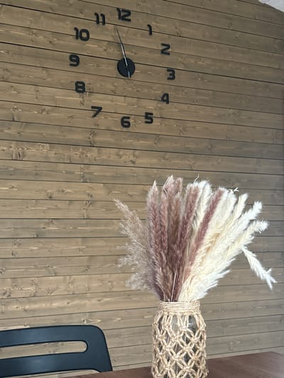 The image shows a wall clock with large, black numbers on a wooden plank wall. The clock hands indicate the time is approximately 12:10. In the foreground, a vase filled with dried pampas grass is placed on a wooden surface. A portion of a black chair is visible in the lower left corner. The setting appears to be indoors. There are no people visible in the image.