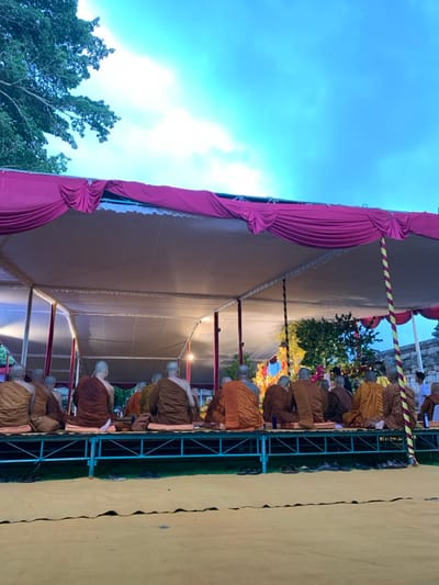 A group of 15 Buddhist monks are seated facing a large, elaborately decorated altar in a large tent structure. The tent has a white fabric roof and pink fabric drapes lining the front and sides. It is located in the city of Mungkid, Indonesia at coordinates -7.605, 110.23. There are a number of people seated on benches in front of the monks, with one person in a bright green shirt and orange pants and another standing, wearing a brown robe. The monks are wearing brown robes and are sitting with their legs crossed. There are a few green plants visible on the ground, as well as a large green tree in the background. The sky is blue with white clouds.