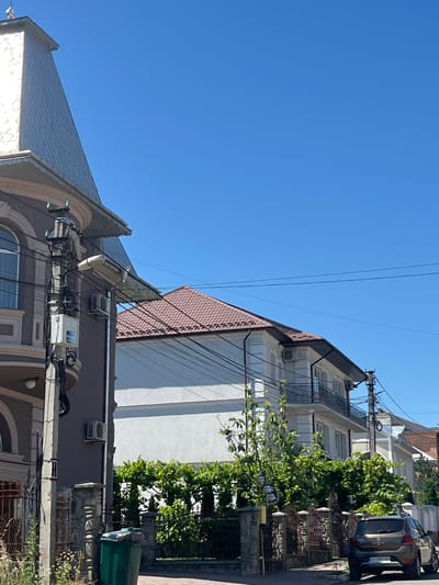 This image, oriented vertically, captures an urban street scene under a clear blue sky, indicating daytime and fair weather. The foreground features a multi-story building with a dark brown facade, adorned with decorative moldings and arched window details. Several air conditioning units are mounted on its exterior. A complex network of electrical wires and utility poles runs across the building's facade, suggesting a residential or mixed-use area.

Behind this building, another structure with a white exterior and a sloped, reddish-brown tiled roof is visible. This building also features balconies and what appear to be air conditioning units. To the right, a dark-colored car is parked on a paved street, with its rear license plate partially visible. Beyond the buildings, dense green foliage suggests the presence of trees and vegetation. Decorative metal fencing and structures are also present, hinting at property boundaries or garden features.

The scene appears to be a quiet street in a European-style town or city, possibly in Chahor, Ukraine, as indicated by the location context. There are no people clearly depicted, but the presence of parked cars and air conditioning units suggests it's an inhabited area. The overall impression is one of everyday urban life on a sunny day. No discernable text other than on the license plate, which is too blurry to read.