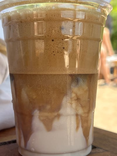 A close-up, eye-level shot shows a clear plastic cup filled with a layered beverage. The top layer is a light brown foam with small bubbles. Below the foam, the drink transitions to a darker brown liquid with ice cubes visible through the condensation on the cup. The bottom layer is a creamy white liquid, with streaks of the darker brown liquid dripping down. The cup is sitting on a wooden surface. In the background, slightly out of focus, a person is visible, and beyond that, green foliage suggests an outdoor setting, potentially a cafe or restaurant. This scene is located in Chernomorets, Bulgaria.