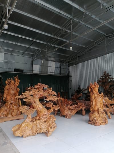 The image depicts a warehouse-like space in Ha Tu Ward, Vietnam, filled with wooden carvings. The space has a corrugated metal roof supported by a metal frame. Inside, numerous intricate wooden sculptures are displayed on a white tiled floor. Prominent among these are a carving of a laughing Buddha figure holding a sack, a tree-like sculpture with detailed foliage, and another standing figure. Other carved wooden items, possibly furniture or decorative pieces, are scattered throughout the room. The light in the warehouse is diffuse, suggesting natural light or fluorescent lighting. The background reveals more of the warehouse structure, including what appears to be a closed metal door.