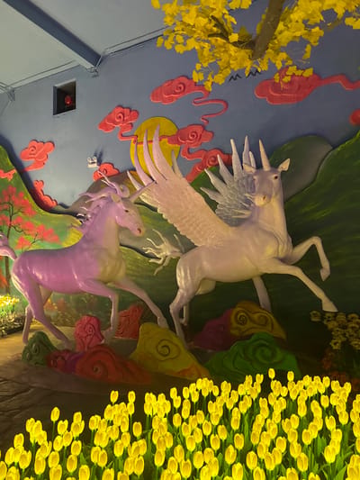 The image depicts a ceiling decoration in what appears to be a tourist attraction or commercial space in Binh Lu, Vietnam. The ceiling features two unicorns: one white with wings, and one purple without wings. The unicorns are set against a backdrop of painted elements, including yellow flowers resembling tulips, yellow leaves on a tree branch, red cloud-like shapes, and green foliage. A square vent with a red interior is visible in the lower left corner of the image. The ceiling's color is primarily blue.