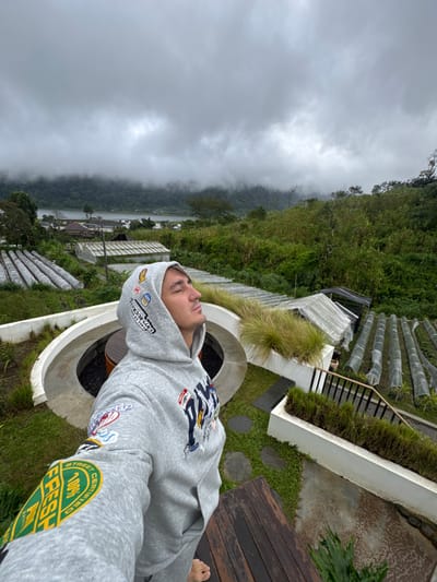 A man lies on his back, wearing a grey hoodie, on what appears to be a patio or deck. The surrounding landscape features a lush, green hillside with terraced structures, possibly for agriculture. A body of water is visible in the distance, partially obscured by heavy mist or fog. The sky is overcast, contributing to a cool and possibly damp atmosphere. The architecture seems modern and minimalist.