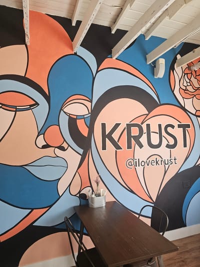 The image shows a section of the interior of "Krust" in Key Largo, Florida, United States of America. The mural on the wall is a colorful, abstract design with the word "KRUST" and the Instagram handle "@ilovekrust" prominently displayed. There is a dark brown table with a black metal frame and a small container with flowers on top. A portion of a wooden staircase is visible on the left side of the image. The overall aesthetic is modern and artistic.