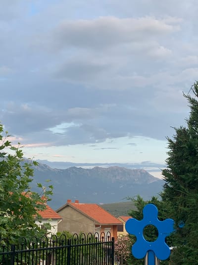 The image depicts a view of the mountains from Radovići, Montenegro. In the foreground, a black metal fence runs horizontally across the bottom of the frame. Behind the fence, there are several houses with orange tile roofs. To the left, a tree with green leaves partially obscures the view. To the right, a large green conifer tree is prominent. A bright blue, flower-shaped object, possibly a playground feature, is visible in the middle right of the image. In the background, a range of mountains stretches across the horizon, shrouded in a hazy, muted light. The sky above is filled with a mix of light and dark clouds.