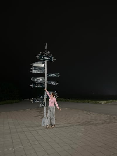 A young woman, wearing a pink long-sleeved shirt and grey pants, is posing next to a signpost in what appears to be an outdoor public space at night. The signpost features multiple directional signs pointing to various cities and their distances, including "ЕЛИЗОВО", "ХАБАРОВСК", "ВЛАДИВОСТОК", "ЛУГАНСК", "КАЛЬЯО", "БЕЛГРАД", "ПЕКИН", "САНКТ-ПЕТЕРБУРГ", "МОСКВА", "Токио", "КРЫМ", "ДОНЕЦК", "МАРКОПОЛЬ", and "Минск". The woman is balancing on one leg with her arms outstretched, seemingly interacting with the signpost. The ground is paved with square tiles, and a strip of grass is visible to the left of the paved area. The background is dark, indicating it is nighttime. The overall scene suggests a playful moment of discovery or a photo opportunity at a location that marks distances to various significant cities. The text on the signpost provides specific location and distance information, with time zone indicators also visible.