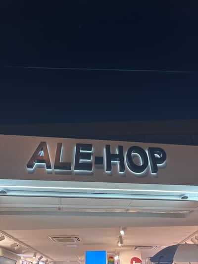The image shows a vertical, illuminated sign that reads "ALE-SHOP" in large, dark, three-dimensional letters. The letters are outlined with a bright white light, creating a halo effect. The sign is mounted on a white vertical surface, likely the exterior of a building or storefront. To the left of the sign is a large, dark blue or black area, possibly a wall or a large pane of glass reflecting the dark exterior. The right side of the image shows a brightly lit interior space, with multiple ceiling lights and track lighting illuminating a white wall. There are some framed objects on the wall and a blue screen is visible in the upper right corner. The lighting suggests it is nighttime or an indoor setting. The scene appears to be a commercial or retail environment. There are no people or specific weather conditions visible.