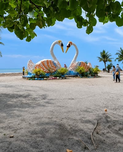 This image captures a scenic view at Kota Pari, Indonesia, featuring a large statue of two white swans with their necks intertwined, forming a heart shape. The swans are adorned with orange and blue patterns, and are placed on a circular platform decorated with potted plants and colorful stones. The statue is situated on a sandy beach, with the calm blue ocean and a clear sky visible in the background. Palm trees are scattered along the shoreline.
In the foreground, a sandy path is visible, with shadows cast from overhead foliage. Several people are present in the scene. On the left, a person in a yellow shirt is standing near the water's edge, appearing to take a photo. Further back, to the right of the swan statue, two women are walking away from the viewer. One woman is wearing a light-colored top and dark pants, carrying a bag over her shoulder, while the other is wearing a darker outfit. The overall atmosphere suggests a peaceful, recreational coastal environment.