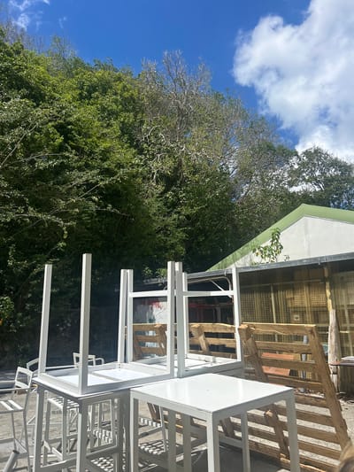 The image shows a daytime scene in Sainte-Anne, France. In the foreground are stacked white metal table frames on a concrete surface. In the background there is a building with a green and white roof and a wire mesh enclosure. A wooden pallet or similar structure leans against the building. Beyond the building, dense green foliage and trees fill the frame, with a blue sky and white clouds visible in the upper left corner. The photograph is taken at an angle.
