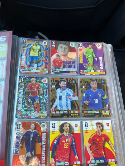 The image shows a binder filled with trading cards, likely football (soccer) cards, arranged in rows. The cards feature portraits of various football players with their names and statistics displayed. Some of the visible players include Vinicius Junior, Joao Neves, Diogo Costa, Lamine Yamal, Lionel Messi, Kylian Mbappe, and Dean Huijsen. The cards have a colorful and shiny design. The binder is open and placed on a dark, textured surface, possibly a bag or upholstery, under artificial lighting. There are no discernible elements that suggest a location in Annalong, United Kingdom; the environment appears to be indoors and generic.