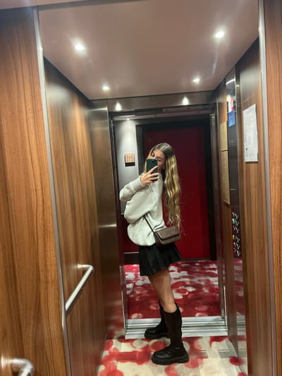A young woman stands in an elevator in Val-d'Isère, France. The woman is wearing a white sweater, a black mini-skirt, and black knee-high boots. She is holding a black purse and looking at her phone which she is holding in her right hand. The elevator walls are made of dark wood paneling. There is a silver handrail on the left-hand wall of the elevator. The elevator floor is covered in a red and black patterned carpet. There is a red door in the back of the elevator which has a silver frame. On the right-hand wall of the elevator, there are several buttons in a vertical row. The elevator is located in a building that has a modern design. The elevator is empty except for the woman. The woman is standing to the left of the center of the elevator and is looking to the right. The woman is facing forward. The woman's hair is long and blonde and is pulled back from her face. The woman's face is visible in the reflection of her phone.