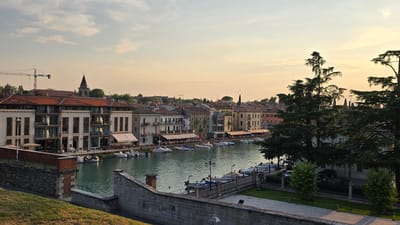 This image depicts a picturesque canal-side scene in Peschiera del Garda, Italy, during what appears to be late afternoon or early evening. The golden hour light bathes the buildings and water in a warm glow, suggesting pleasant weather.

The main subjects are the buildings lining the canal, characterized by their terracotta roofs and pastel-colored facades. Many of these buildings feature awnings shading outdoor seating areas where numerous people are visible, seemingly enjoying meals or drinks at restaurants and cafes. Small boats are moored along the canal, some with people aboard.

The canal itself is a prominent element, its tranquil, turquoise waters reflecting the sky and surrounding architecture. On the right side of the image, large, mature trees with dark green foliage create a natural contrast with the urban landscape. In the background, a church steeple and a construction crane hint at the town's blend of historical and modern elements.

The scene conveys a sense of relaxed activity and leisure, typical of a European town square or waterfront promenade. The overall mood is peaceful and inviting. No discernible text is visible in the image.