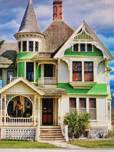 The image is a photograph of a large, multi-story house with a distinctive architectural style. The house is primarily white with vibrant green accents. It features a conical turret with a shingled roof, multiple windows of various sizes and shapes, and a prominent front porch with a decorative railing and a rounded opening. A brick chimney rises from the roof. The house is surrounded by a lawn and appears to be in a suburban or rural setting, with a blue sky and scattered clouds visible in the background. There are no people visible in the image. The provided image does not contain any visual information that would allow for its location to be identified as Sluck, Belarus.