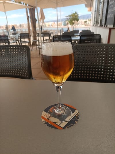 A goblet-style glass containing an amber liquid topped with a white frothy head, exhibiting condensation on its exterior, rests on a circular coaster featuring an abstract geometric design in orange, blue, and white. This setup is on a flat, light gray table. The scene is an outdoor patio or terrace in a coastal area of Agüimes, Spain. In the mid-ground, numerous black, perforated plastic chairs and tables are visible, some positioned under large white patio umbrellas. Beyond the seating area, a waterfront is apparent, leading to a body of water. Distant buildings line the shore, backed by mountains under a clear blue sky with minimal cloud cover. Several indistinct human figures are sparsely distributed in the distant background. The overall lighting indicates a bright, sunny daytime environment.