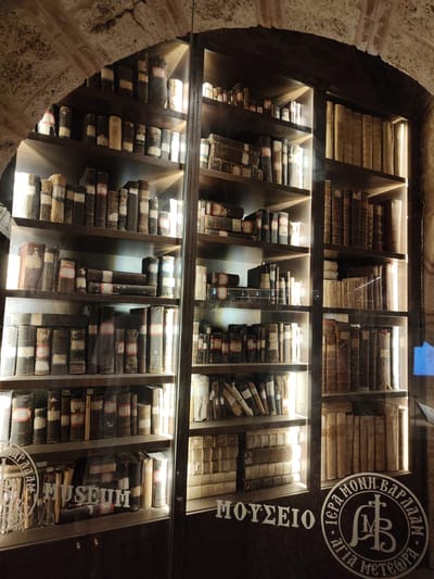 The image shows an interior view of a glass-enclosed bookshelf filled with numerous old books, located within the Holy Monastery of Great Meteoron in Kalambaka, Greece. The monastery is a UNESCO World Heritage site renowned for its historical and religious significance, situated atop towering rock formations. The bookshelf is made of dark wood with multiple shelves, illuminated from behind, creating a warm glow that highlights the spines of the ancient volumes. Many of the books appear to be leather-bound, with some having visible labels or markings. The environment suggests a museum or archival setting within the monastery, preserving these historical texts. Visible text in Greek on the glass includes "ΜΟΥΣΕΙΟ" (Museum) and a circular emblem with text relating to "ΙΕΡΑ ΜΟΝΗ ΒΑΡΛΑΑΜ" (Holy Monastery of Varlaam), indicating the location's connection to the Meteora monastic community. The architectural elements of the monastery, such as the arched stone ceiling visible in the upper left, are also present. There are no people or actions depicted in the image; it is a still scene focused on the display of books.
