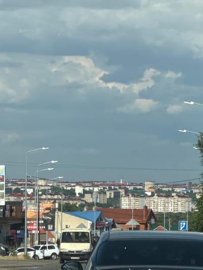 This is a sideways, eye-level shot of a street scene in Dorozhny, Russia, likely taken from inside a vehicle. The image is dominated by a cloudy sky on the left and a city street with buildings and vehicles on the right. The time of day appears to be daytime, given the brightness, though the heavy cloud cover suggests it might be overcast or approaching rain.
On the right side of the frame, a variety of buildings can be seen, ranging from multi-story apartment complexes with cream-colored facades to smaller, residential structures with red-tiled roofs. There is a prominent white panel van parked on the street, partially covered by a blue tarp. Several cars are also visible, including a white hatchback. A traffic sign indicating a mandatory turn is present. Streetlights on poles line the road.
There are several billboards and signs with Russian text visible along the street. One billboard has large, colorful graphics and what appears to be a phone number. Another sign includes the Cyrillic words "РОССИЙСКИЙ" and "НАРОДНЫЙ", suggesting a public or national theme.
The overall impression is that of a typical urban environment on a cloudy day. There are no people clearly visible in the frame, but the presence of vehicles and buildings indicates human activity and habitation. The composition is dynamic due to the sideways perspective, making it feel like a candid snapshot of everyday life.