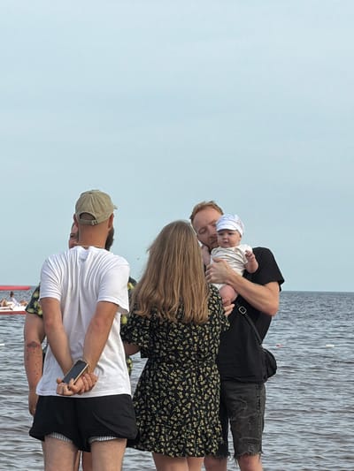 This image depicts a group of people by a body of water, likely a lake or sea, under a clear sky. The setting appears to be outdoors, possibly a waterfront recreational area in Ondvor, Russia. The time of day is likely daytime, suggested by the bright, even lighting.

The central subjects are four individuals: a baby being held by a man, a woman with long blonde hair standing next to them, and a man in a white t-shirt and black shorts in the foreground. The baby is wearing a white onesie and a light-colored cap. The man holding the baby is wearing a black t-shirt and distressed denim shorts. The woman is wearing a dark floral print dress. The man in the foreground is wearing a white t-shirt, black shorts, and a beige baseball cap, and he is holding a smartphone.

The group seems to be engaged in a casual gathering. The interaction between the man holding the baby and the woman suggests a familial or close relationship. The man in the foreground appears to be observing the scene or perhaps waiting. The overall mood is relaxed and serene.

Notable details include the textured water in the background, which suggests a calm body of water with some ripple. The clothing of the individuals indicates a warm weather day. The presence of a bag slung over the shoulder of the man holding the baby adds to the casual outdoor atmosphere. There is no visible text in the image.