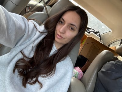 A young adult female is positioned in the front passenger seat of a vehicle, capturing a selfie. She has long, dark brown hair and is wearing a light gray crew-neck sweatshirt. Her right arm is extended, holding the camera device. The vehicle's interior features light tan or beige upholstered seats. The rear passenger compartment is occupied by various items, including a large brown cardboard box, possibly for an electronic device or appliance, and a stack of items wrapped in pink and white plastic, consistent with baby diapers, visible on the floor or seat below the box. Additional indistinct items and fabric are present, suggesting the vehicle is packed. The scene occurs within a vehicle in the city of Golyamo Belovo, Bulgaria.