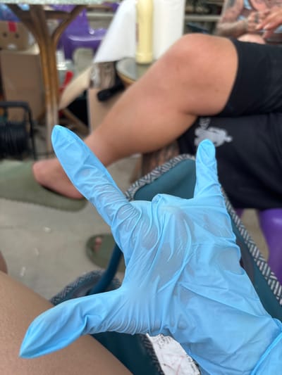 The image displays an indoor or semi-outdoor scene located in Kostievo, Bulgaria, captured from a low, slightly downward perspective. In the foreground, a light-skinned left hand is prominently featured, encased in a light blue disposable glove. The gloved hand is performing a gesture where the index finger, pinky finger, and thumb are extended upwards, while the middle and ring fingers are curled inward. Irregular patterns of moisture or residue are visible on the surface of the glove. A small section of a white paper containing indistinct black text is partially visible beneath the gloved hand.
In the midground, the bent right leg of another individual, exhibiting a light to medium skin tone, is visible. This person is wearing black mesh-textured shorts or athletic wear. Further in the background, the upper torso and a portion of a tattooed arm belonging to this individual are present, appearing out of focus. The person's left foot, adorned with an olive-green slide sandal, is visible resting on a light-colored floor.
The immediate environment includes a teal-colored portable chair or seat with a grey and white patterned edge, which is supporting the arm of the gloved hand. In the blurred background, several objects are discernible: a light brown table with a transparent surface, a partial purple chair or stool, a tall yellow bottle, and a roll of white paper towels positioned on an elevated surface. A coiled dark object, possibly a cable or hose, is situated on the ground in the lower left portion of the frame. The floor throughout the visible area is light-colored, consistent with concrete or tile.