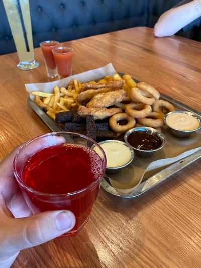 A close-up, eye-level shot captures a hand holding a glass of red beverage with frothy bubbles, positioned in the foreground. Behind it, a metal tray laden with a variety of appetizers rests on a wooden table. The tray contains French fries, fried chicken strips, onion rings, and dark croutons. Three small bowls of dipping sauces—two white and one dark—are arranged on the tray. In the background, two shot glasses filled with a red liquid and a tall glass with a yellow beverage and a straw are visible. The setting appears to be a restaurant or bar. The image location is not specified in the provided information.