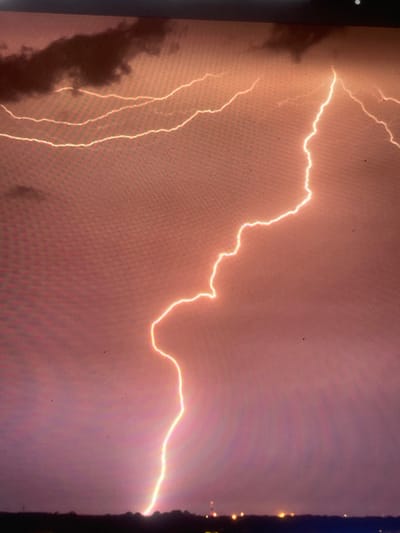 The image displays a prominent lightning strike illuminating a stormy night sky over Zelenyi Hai, Ukraine. A singular, intensely bright, and highly jagged lightning bolt originates from the upper-right cloud mass and descends diagonally, connecting with the distant ground line in the lower-center. Multiple thinner, horizontal lightning flashes are embedded within the dark, dense cloud cover occupying the upper-left quadrant. The sky is characterized by deep reddish-orange to purple coloration, indicative of the atmospheric conditions during the electrical discharge. A dark, silhouetted landmass, appearing as a distant treeline or low hills, forms the horizon across the lower portion of the frame. Scattered small, yellow-orange artificial lights are visible below this horizon, suggesting distant infrastructure or a settlement. No individuals or specific objects are identifiable on the landmass. The overall image exhibits a pixel grid pattern, consistent with a photograph taken of a digital screen, and includes minor dust particles or screen imperfections.
