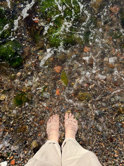 The image shows a top-down view of a person's feet submerged in shallow, clear water on a pebble beach. The water is gently lapping, creating small waves and foam around the feet. The pebbles on the beach are varied in color and size, with some small pieces of green seaweed visible. The person is wearing light-colored pants that extend down to their ankles. The lighting suggests it is daytime. The specific location of Üsküdar, Turkey, cannot be determined from the visual information provided in the image.