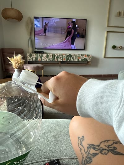 In a living room, likely in San Diego, California, a person's hand is seen holding the cap of a large plastic water bottle. The bottle is partially filled with water and has condensation on its exterior. The person is wearing a light blue sweatshirt. In the background, a television screen displays a video of two women practicing a dance routine in a studio. A decorative cabinet with a cactus pattern sits beneath the television, and a vase with dried reeds is placed to the left of the TV. Two framed pictures hang on the wall to the right. The scene appears to be a casual moment captured at home, possibly during leisure time.