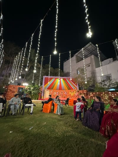 Outdoor nighttime event featuring string lights. A vibrant backdrop, presumably for a ceremony or stage, is the focal point. People are gathered on the lawn, some seated and others standing. Several individuals are dressed in formal attire or traditional garments. The event seems festive, set against a backdrop of a building.