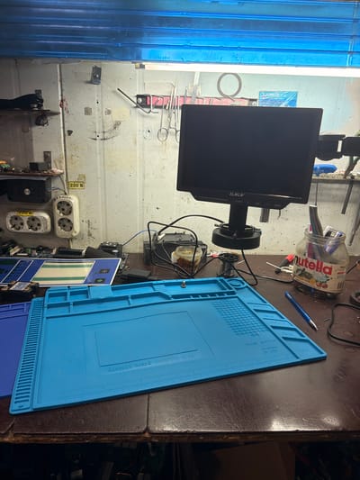 The image shows a workbench in what appears to be a repair or electronics workshop, possibly located in Yermolino, Russia. The workbench is cluttered with tools and electronic components. Prominently displayed is a black monitor mounted on a stand, likely part of a microscope setup. In the foreground, a bright blue mat with grids and labels for "Parts" and "magnetic" is laid out, suggesting a workspace for assembling or repairing small electronic items. Various tools, including screwdrivers, soldering equipment, and what look like tweezers or forceps, are visible on shelves and the workbench. Several electrical outlets are mounted on the wall behind the workbench, with one labeled "220 B" and another bearing Cyrillic text. A jar of Nutella is repurposed as a container for pens and other small tools. The overall environment suggests a functional, though somewhat disorganized, workspace.
