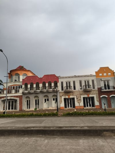 The image depicts a street scene in Babakan Madang, Indonesia, showcasing a row of distinct, multi-story buildings under an overcast sky. A tall, black streetlight pole curves into the frame from the left. The buildings exhibit varied architectural styles and colors, including orange, red, white, grey, and light blue accents.
The leftmost building features a large, rounded orange roof and prominent arched windows. Adjacent to it, a building with a red upper section displays multiple white arches above ornate black-railed balconies. The central structures are predominantly white and grey, adorned with tall windows and smaller balconies. The building on the far right has a striking orange, stepped gable roofline and two light blue arched doors at ground level. Some facades show textured finishes like brickwork or stone.
In the foreground, a concrete road stretches across the frame, displaying slight motion blur, suggesting the photo was taken from a moving vehicle. A curb and a strip of low green vegetation separate the road from the buildings. On the right, a person in a light shirt walks along the sidewalk, while another figure is partially visible near the base of the lamppost on the left. The grey, cloudy sky casts a soft, even light over the entire scene. There is no visible text within the image.