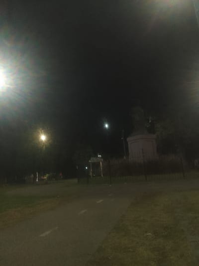 A dark, nighttime scene captures a park setting. A paved path stretches across the lower half of the image, bordered by grassy areas. In the distance, a monument or statue stands prominently, illuminated by a faint moon or light source. Streetlights cast a bright glare on the left and right sides of the frame. Fences and dense shrubbery line the perimeter of the park area. The overall atmosphere is shadowy and quiet.