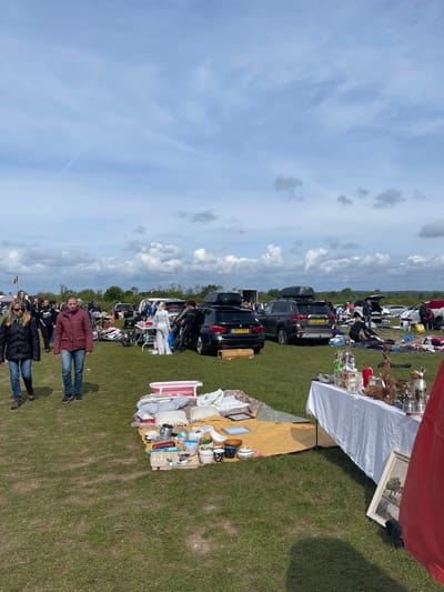 The image depicts a car boot sale in Sevenoaks, United Kingdom. In the foreground, two women stand on the grassy field. Cars are parked in rows, forming the backdrop to the sale. Tables display an assortment of items for sale, including ornaments, crockery, and framed pictures. People are browsing and interacting with the vendors. The sky is blue with scattered clouds.