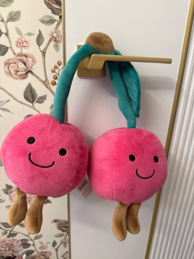 Two pink plush cherry toys with black embroidered smiling faces and brown legs hang from a gold-colored door handle. The toys have green stems and leaves. The background features a wall with floral wallpaper on the left and a white fluted panel on the right. The image appears to be taken indoors in a residential setting in Razdolnoye, Russia.