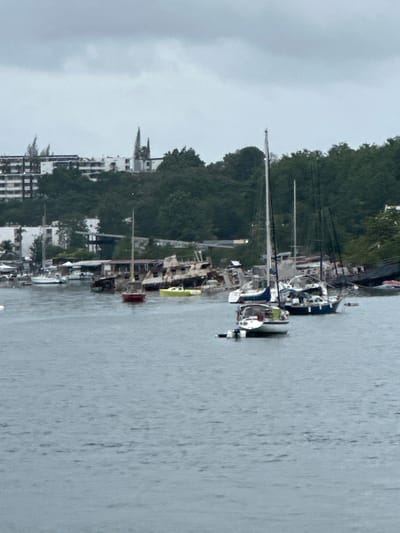 The image depicts a harbor scene in Pointe-à-Pitre, France. The sky is overcast and gray. The water in the foreground is choppy. Several sailboats are docked or anchored in the water, with their masts visible. In the mid-ground, there are various moored vessels, some appearing to be in a state of disrepair, including a partially submerged boat and a bright yellow boat. Further back, on the shore, there are buildings, including a multi-story white building with many windows and a lush green forested hillside. Two pointed spires rise above the trees on the hill, possibly part of a church or other structure. The overall impression is a working harbor with a mix of active and derelict boats, set against a backdrop of urban development and natural landscape.