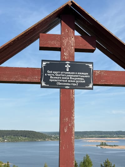 A wooden cross stands against a blue sky backdrop with a view of a body of water and greenery. Attached to the cross is a black plaque with white lettering in Russian. The text on the plaque reads: "This cross is installed and consecrated in honor of the Holy Equal-to-the-Apostles Great Prince Vladimir, the enlightener of the Russian land (Bessonov Mountain)". This indicates the location is likely Elkino, Russia.