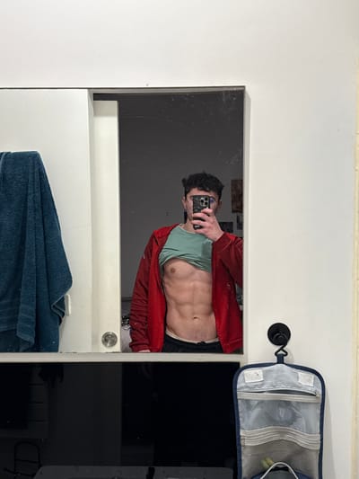 A man is reflected in a mirror, taking a selfie with his phone. He is wearing a red jacket over a light green shirt and has his stomach exposed. A dark blue towel hangs below the mirror, and a black countertop with a hanging toiletry bag is visible to the right. The surrounding wall is white. The mirror shows smudges and reflections.