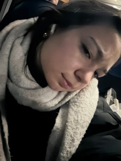 A young woman with dark hair and subtle winged eyeliner looks towards the viewer with a slight frown. She is wrapped in a thick, textured white scarf. She appears to be wearing a dark-colored shirt or jacket. She wears a small earring in her left ear. A portion of what seems to be a garment or jacket is visible in the upper right corner of the frame. The image has low lighting.