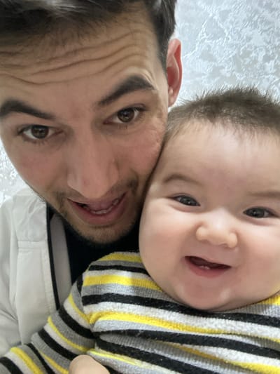 The scene depicts a close-up indoor portrait of an adult male and an infant, taken in Yangiiul, Uzbekistan. The adult male, positioned on the left, has dark hair, brown eyes, and light facial hair. He is wearing a black turtleneck under a white jacket, facing the camera with a smiling expression, his mouth slightly open. The infant, on the right, has a round face, dark eyes, and sparse dark hair. The infant is wearing a long-sleeved shirt with horizontal black, yellow, and grey stripes, and is smiling widely at the camera, revealing two lower front teeth. The adult appears to be holding or embracing the infant. The background consists of a light-colored wall with a subtle textured pattern.