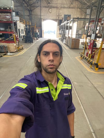 A man is standing in a large, industrial warehouse-like building in Sáenz Peña, Argentina. The building has a high ceiling with exposed brick and metal support beams. There is natural light coming from a large opening at the far end of the building. The man is wearing a purple and lime-green work uniform with the word "Emova" on the chest. He is looking at the camera. The floor is concrete with yellow lane markings. Shelving units filled with various items are visible along the sides of the building. There are safety barriers constructed of yellow and black metal tubing. A clock is visible on the upper wall near the back of the warehouse.
