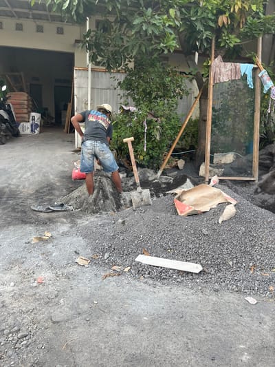 In Kedungkendo, Indonesia, a construction worker wearing a dark t-shirt, denim shorts, and a light-colored cap is partially submerged in a pile of construction gravel. He is actively working with a shovel, digging into the gravel pile. A red bag, possibly containing cement or similar material, is visible near him. A pair of dark flip-flops rests on the ground to his left. In the background, a building with an open garage and stacked bags of construction material, including "WHITE SANDCOAT," are present. A motorcycle is parked to the left. To the right, a wooden frame with mesh and some clothing hanging on it stands near a large tree with lush green foliage. Another pile of gravel and some torn paper bags are also visible in the scene.