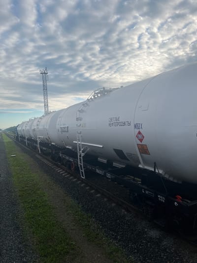 The image shows a long train composed of white tank cars parked on a railway track. The train is positioned on the right side of the frame, extending from the foreground towards the background. The tank cars are cylindrical and white, with various markings and labels visible on their sides. Some of these markings are in Cyrillic script, including what appears to be "СВЕТЛЫЕ НЕФТЕПРОДУКТЫ" and "МТБЗ Х". There are also hazard symbols visible, including a red diamond with "AK 301" and an orange rectangular placard with "33" and "2396". The train is situated on a gravel ballast track, with green grass growing alongside the tracks on the left. In the background, a tall lattice tower, possibly a communication or observation tower, stands against a cloudy sky. The sky is filled with scattered, elongated clouds, some of which are illuminated by the sun. The overall scene suggests an industrial or logistical setting related to the transportation of petroleum products in Kemuly, Russia.